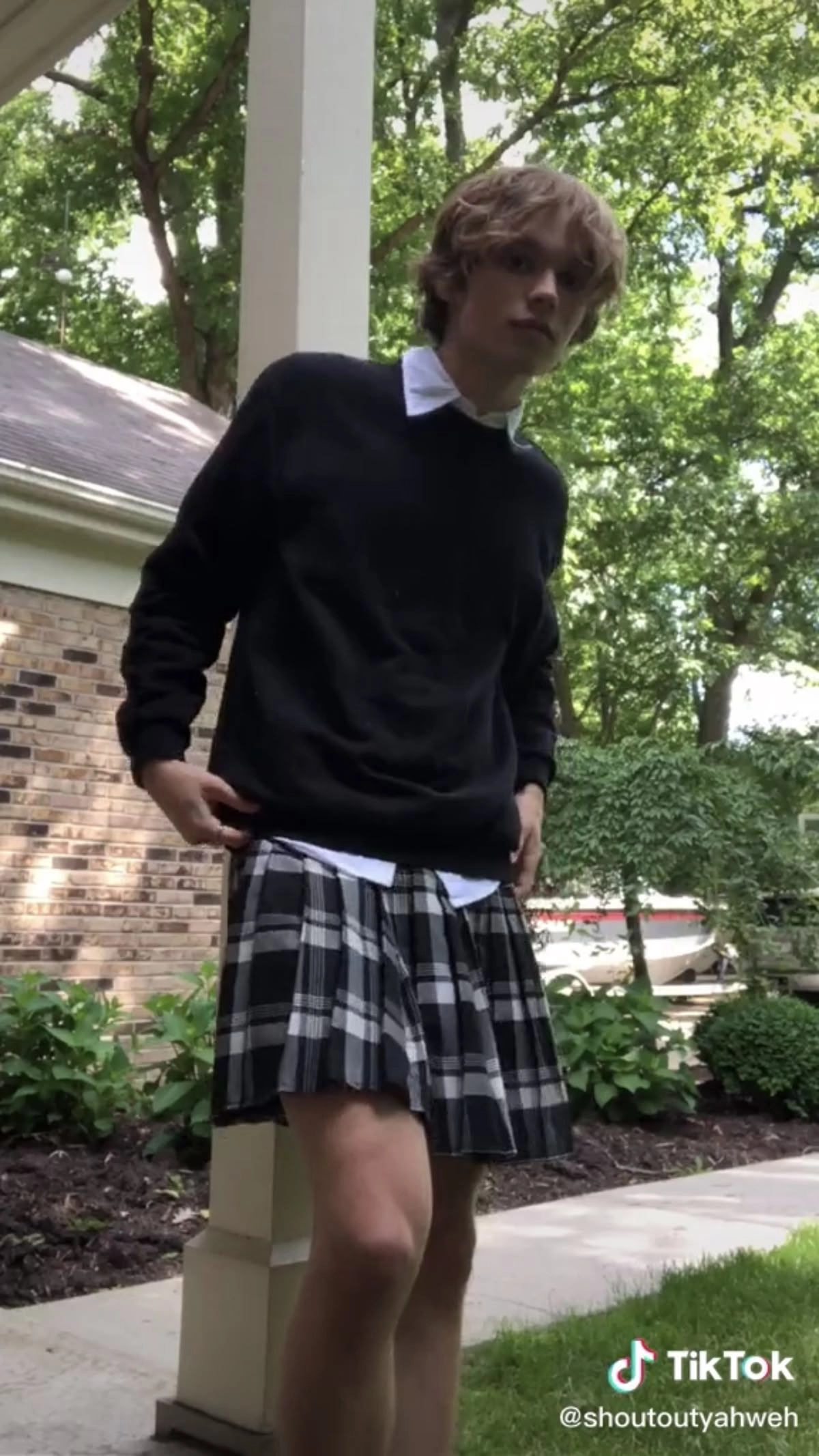A man in a black sweater and plaid skirt standing on a sidewalk.