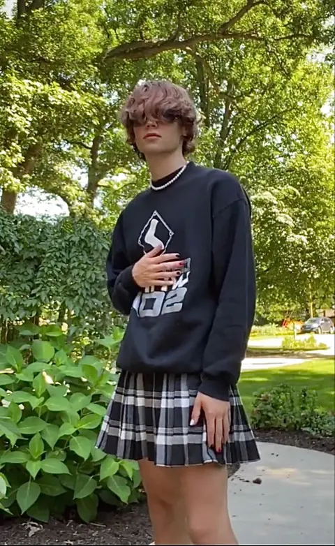 A young man in a black sweatshirt and plaid skirt posing for a picture.