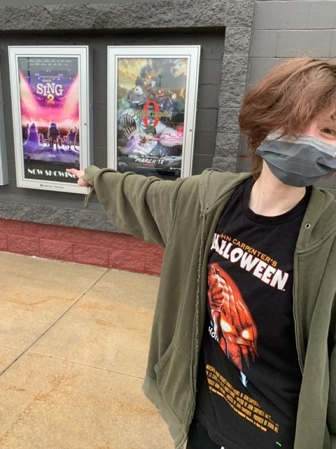 A person wearing a mask standing in front of a wall with posters.